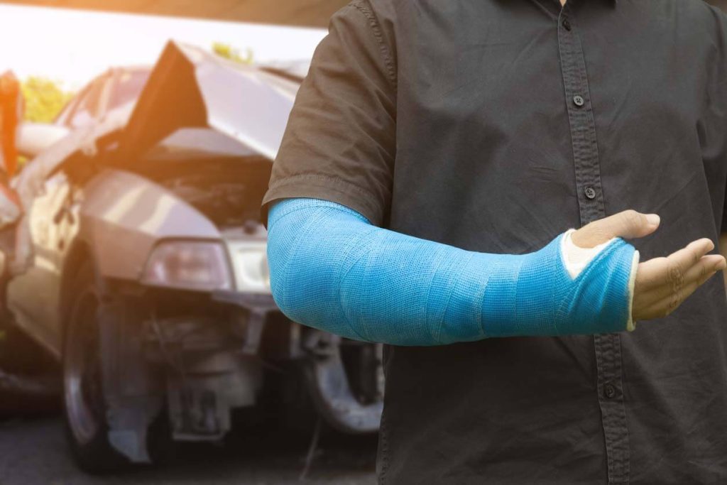 a person with a blue cast on his arm with a wrecked car behind him after he got compensation with the help of his car accident lawyer 